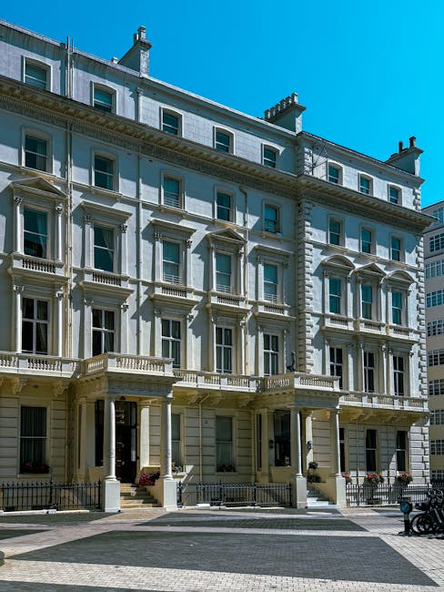 A street scene featuring a large, white, multi-story building with numerous windows and a decorative black railing on the first-floor balcony, situated in a London neighbourhood. The area includes pav